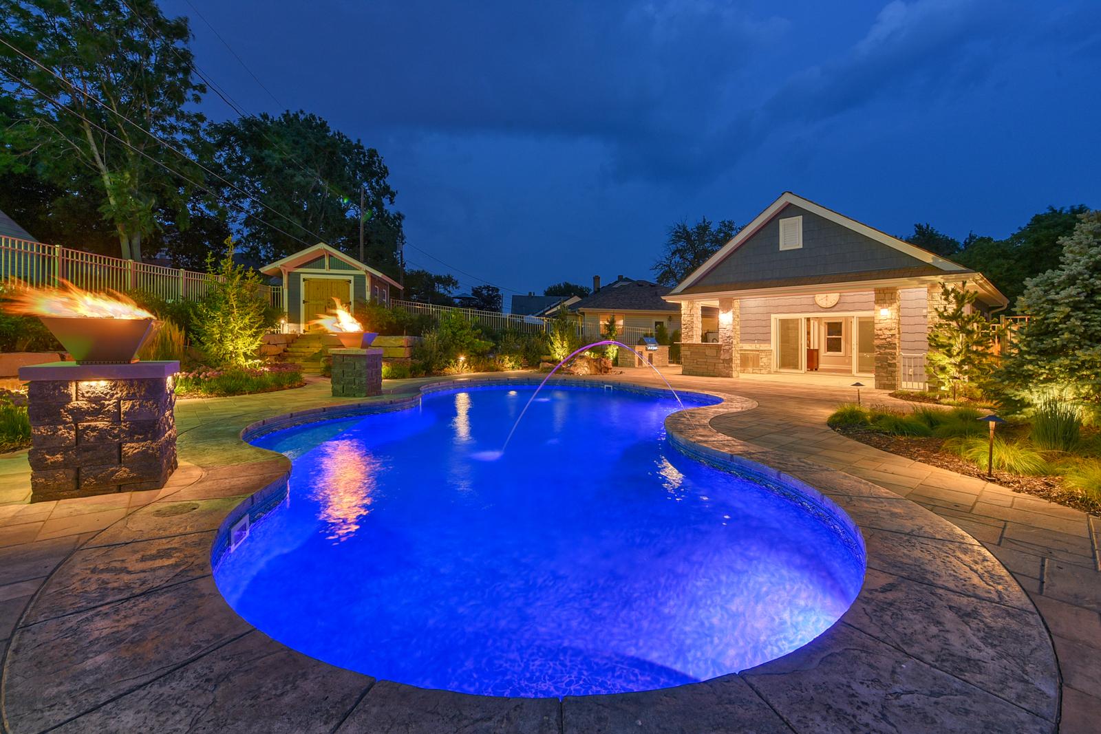 Stunning illuminated pool area featuring fire bowls and elegant landscape lighting creates the perfect evening atmosphere.