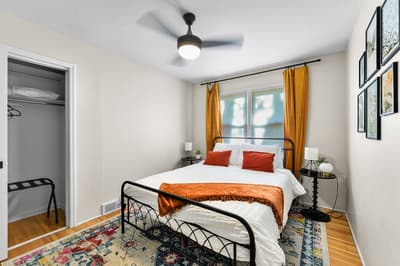 Your cozy bedroom features a comfortable bed with vibrant orange accents, overhead fan for comfort, and warm natural light streaming through golden curtains.