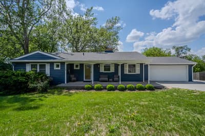 Charming blue ranch-style home with attached garage, surrounded by mature trees and manicured landscaping in a peaceful neighborhood setting.