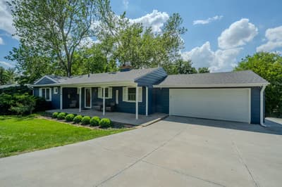 Charming single-story home with blue siding and white trim, featuring a spacious driveway and surrounded by mature trees in a quiet residential neighborhood.