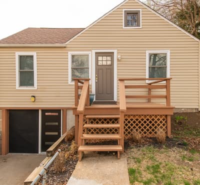 Charming tan-sided home features a welcoming wooden deck entrance with clean white trim and multiple windows throughout.