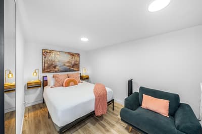 Relax in your stylish bedroom featuring crisp linens, warm coral accents, and a cozy lounge chair perfect for unwinding after exploring the city.
