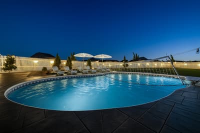 Illuminated pool area with lounge chairs and umbrellas under evening sky, surrounded by modern fencing.
