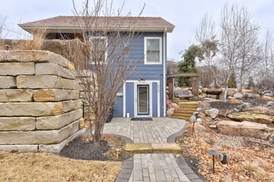 A charming blue cottage nestled among stone walls and mature trees, featuring a welcoming paved entrance walkway.