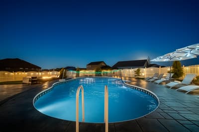 Evening pool area with illuminated blue water surrounded by lounge chairs and residential neighborhood backdrop under deep blue sky.