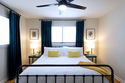 Drift off peacefully in your elegant bedroom featuring crisp white linens, stylish navy curtains, and gentle ceiling fan for perfect comfort.